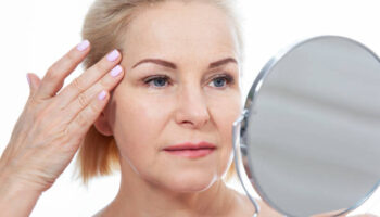 Happy middle aged woman model touching face skin looking in mirror.