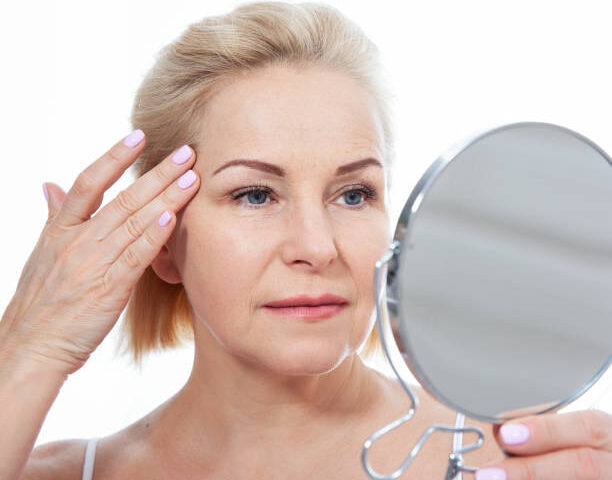 Happy middle aged woman model touching face skin looking in mirror.