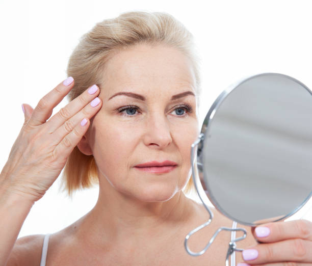 Happy middle aged woman model touching face skin looking in mirror.